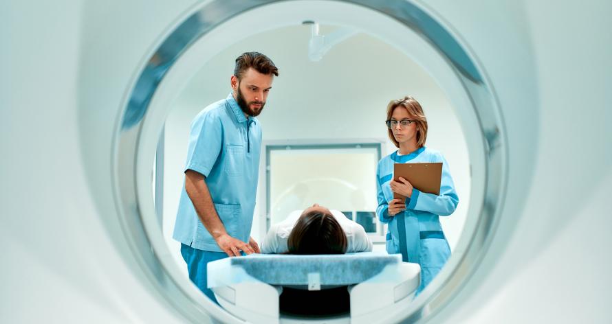 View from inside an MRI scanner showing a patient lying on the table, with two medical professionals in blue scrubs—one holding a clipboard—standing nearby, preparing for cardiac imaging services UK. View from inside an MRI scanner showing a patient lying on the table, with two medical professionals in blue scrubs—one holding a clipboard—standing nearby, preparing for cardiac imaging services UK.